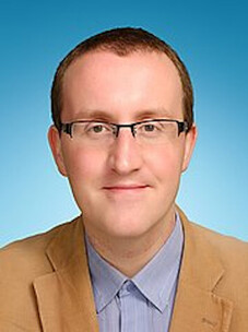 René Meyer mit Brille und beigem Sakko vor hellblauem Hintergrund.